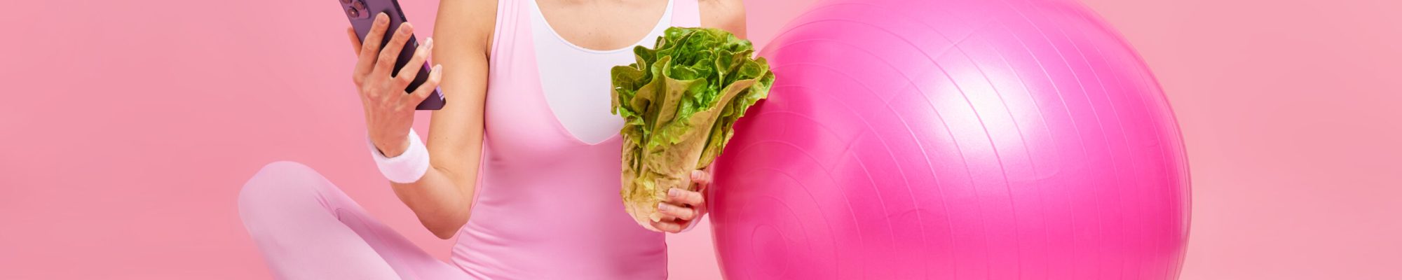 Joyful fitness woman holds mobile phone checks how much calories she burned during workout leads active healthy lifestyle eats greenery keeps to diet sits on mat with sport equipment around.